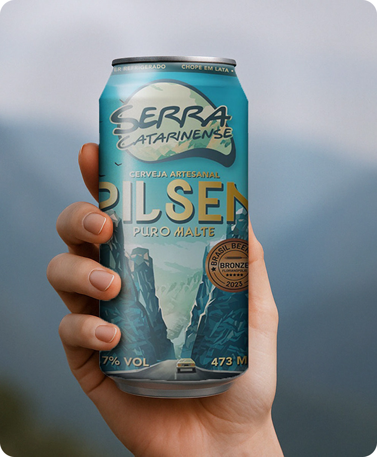 Hand holding a Serra Catarinense Pilsen can, illustrated with a mountain road landscape.